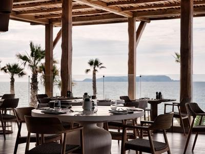Open hotelruimte met tafels en stoelen met uitzicht op palmbomen en de zee.