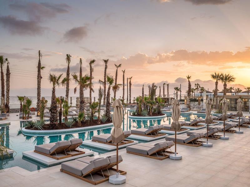Espace piscine luxueux avec chaises longues et palmiers au coucher du soleil au bord de la mer.