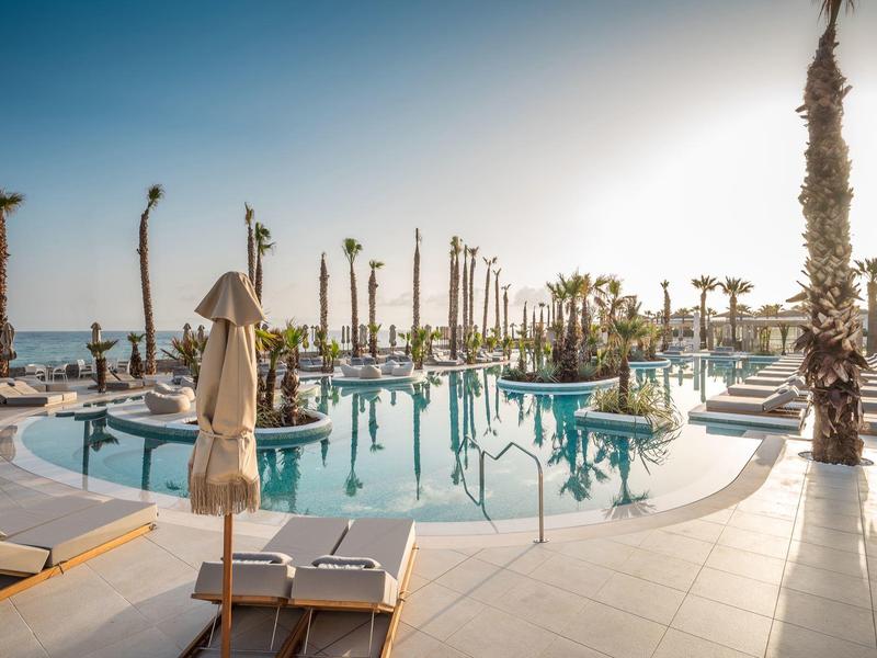 Grande piscine avec palmiers et transats au bord de la mer sous un ciel dégagé.