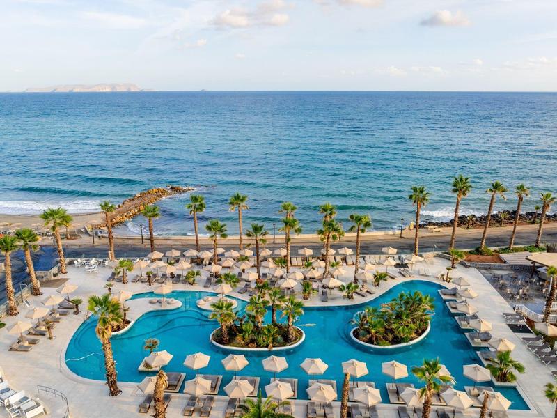 Grande piscine en bord de mer avec palmiers et parasols près de la plage.