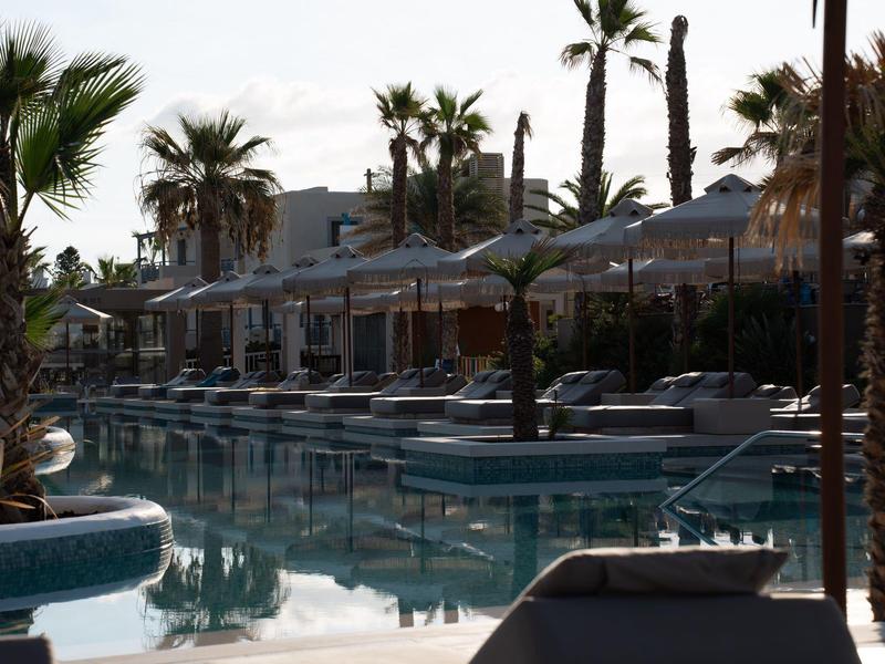 Zone de piscine d'un hôtel avec chaises longues, parasols et palmiers au coucher du soleil.
