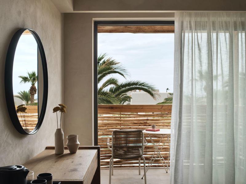 Chambre d'hôtel lumineuse avec meubles en bois, miroir rond et vue sur balcon avec palmiers.