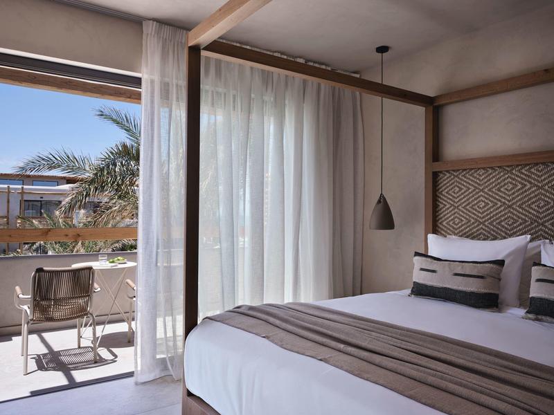 Chambre d'hôtel lumineuse avec grand lit, rideaux et balcon avec chaises et vue sur les palmiers.