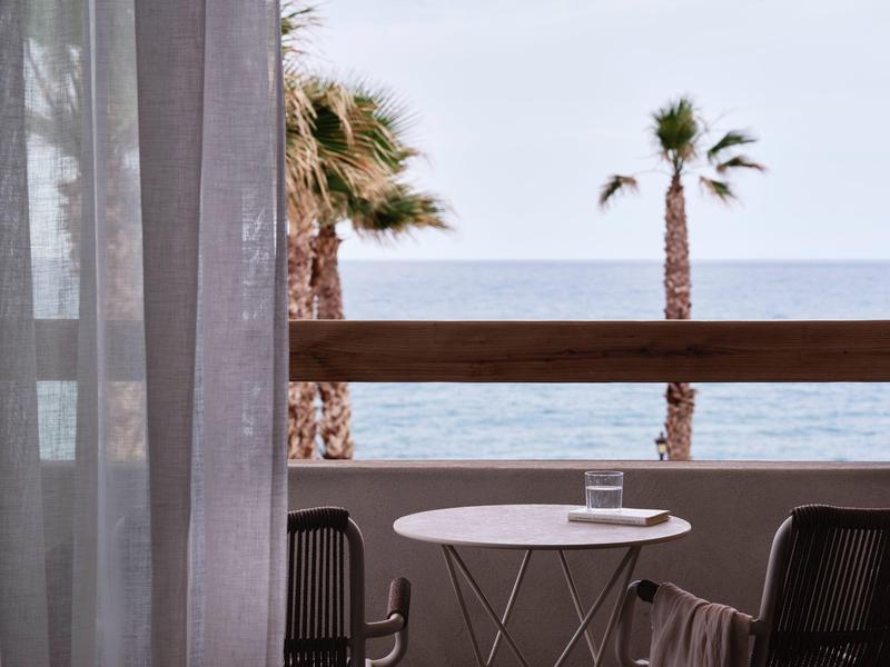 Balcon avec petite table ronde, deux chaises, vue sur des palmiers et la mer en arrière-plan.