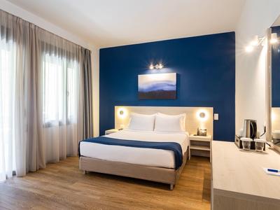 Schlichtes Hotelzimmer mit Doppelbett, blauer Akzentwand, Holzboden und großem Fenster.