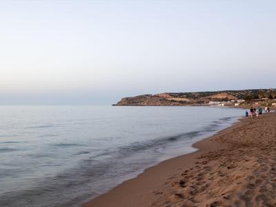 Sandstrand mit ruhiger See, weiter Himmel und einem Felsenabschnitt am Horizont bei Dämmerung.