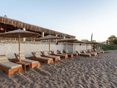 Mehrere Sonnenliegen mit Holzrahmen und beigen Polstern stehen auf sandigem Boden unter Sonnenschirmen.