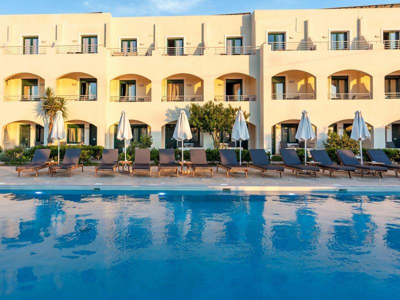 Moderner gelber Hotelkomplex mit blauen Pool und Sonnenliegen bei klarem Himmel.