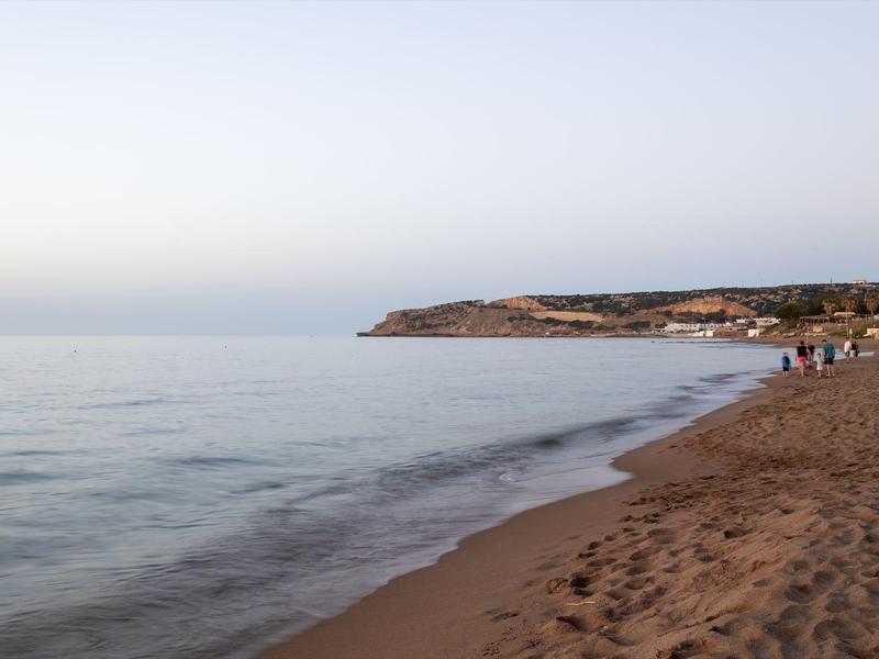 Sandstrand mit ruhiger See, weiter Himmel und einem Felsenabschnitt am Horizont bei Dämmerung.