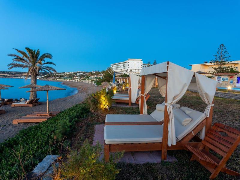 Strand mit weißen Sonnenschirmen und Holzbänken, blaues Meer und klarer Himmel bei Sonnenuntergang.