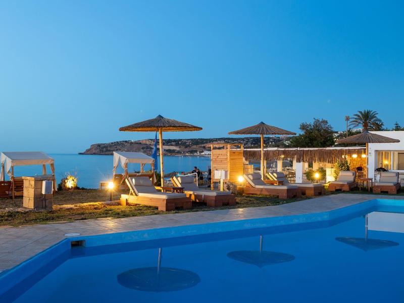 Abendstimmung mit beleuchtetem Pool, Liegestühlen, Sonnenschirmen und Meer im Hintergrund.