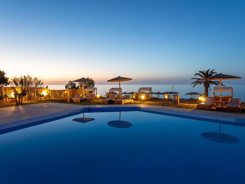 Pool mit klarem Wasser, Liegestühlen und Sonnenschirmen bei Sonnenuntergang am Meer.