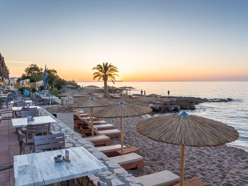 Strandpromenade mit Tischen, Stühlen, Sonnenschirmen und Palmen bei Sonnenuntergang am Meer.