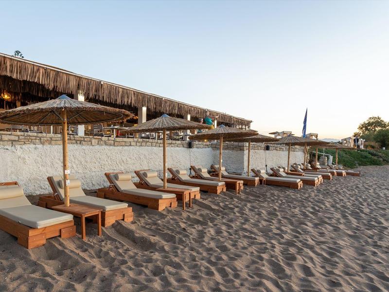Mehrere Sonnenliegen mit Holzrahmen und beigen Polstern stehen auf sandigem Boden unter Sonnenschirmen.