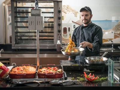 Mann in Kochkleidung serviert frisch zubereitetes Essen in einer Küche mit Grill und Pfannen.