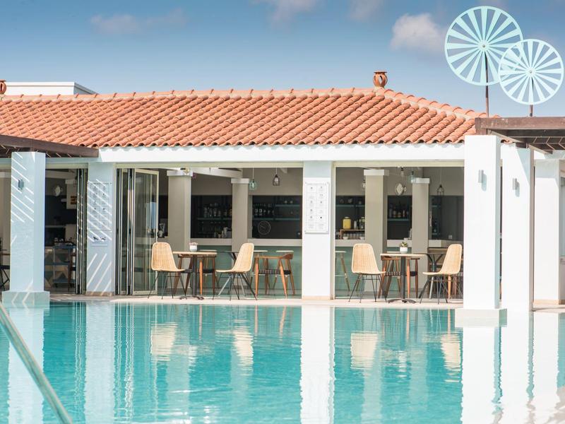 Piscina exterior moderna con agua azul clara y edificio blanco anexo con techo de terracota.