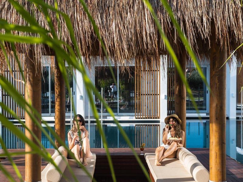 Dos mujeres descansan en tumbonas bajo un techo de paja en una pasarela de madera sobre el agua.