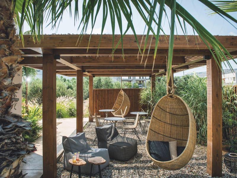 Área exterior acogedora con silla colgante de mimbre y techo de madera en un jardín con palmeras.