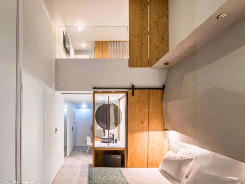 Habitación de hotel moderna con elementos de madera y blancos, espejo y puerta corredera en la entrada.