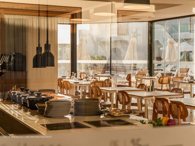 Comedor luminoso de hotel con mesas y sillas de madera y área de buffet con lámparas colgantes.