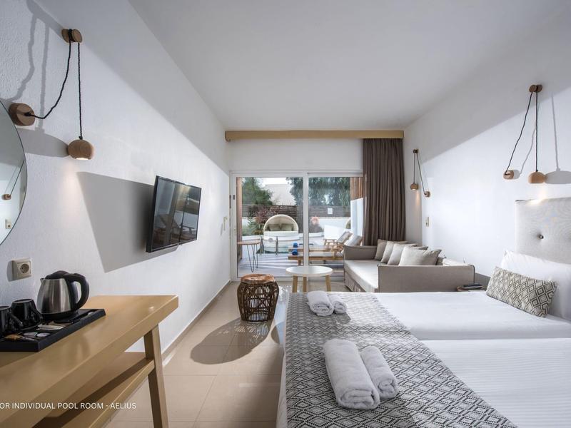 Habitación de hotel luminosa con cama doble, sofá, TV, espejo y lámparas modernas.