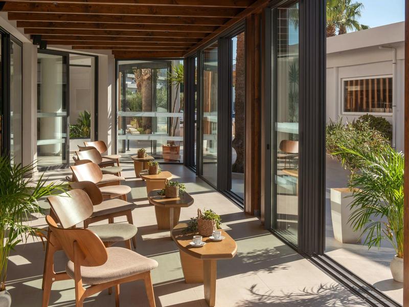 Balcone luminoso con sedie in legno e piccoli tavoli, circondato da finestre in vetro.