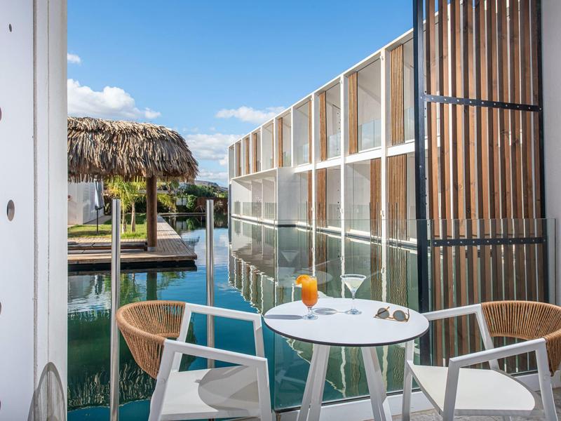 Balcone con piccolo tavolo, due sedie e bevande che si affaccia sull'acqua e ambiente tropicale.