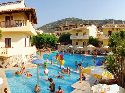 Schwimmbad mit vielen Menschen, umgeben von gelben Gebäuden und grünen Hügeln im Sonnenschein.