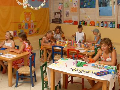 Kinder sitzen an Tischen in einem Raum, der mit bunten Bildern und Bastelmaterialien dekoriert ist.