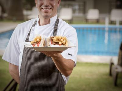 Kelner w białej koszuli i fartuchu trzyma tacę z burgerami i frytkami przy basenie.