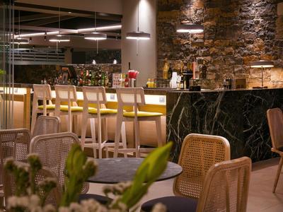 Nowoczesny bar hotelowy z wygodnymi krzesłami, wysokimi stolikami i kamienną ścianą w tle.