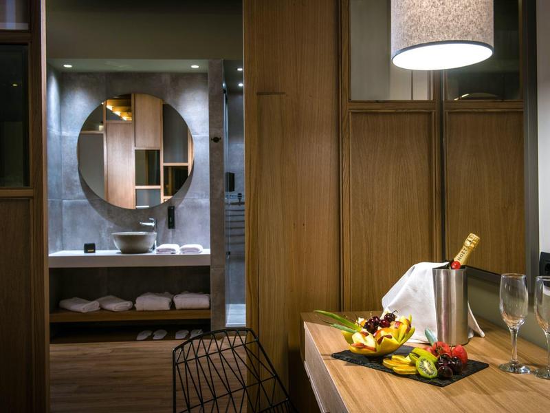 Chambre d'hôtel moderne avec murs en bois, table avec vin et fruits, salle de bain en arrière-plan.