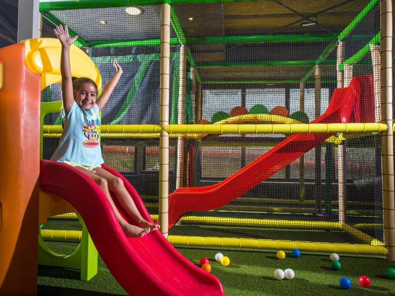 Enfant joue sur un toboggan rouge dans une aire de jeux intérieure colorée avec structure d'escalade et balles.
