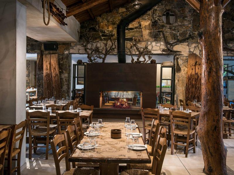 Restaurant rustique avec tables en bois, chaises et cheminée centrale aux murs en pierre.