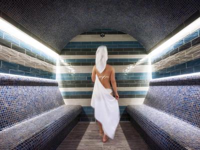 Woman in towel walking in steam room with mosaic walls and benches.