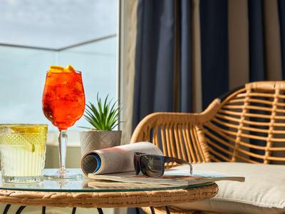 Due bevande su un tavolo con un giornale accanto a una sedia in vimini e tende sullo sfondo.