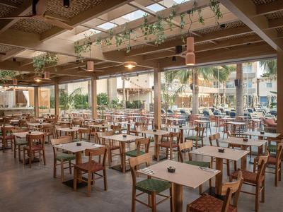 Großes, offenes Restaurant mit vielen Holztischen und -stühlen unter einem Glasdach mit Pflanzen.