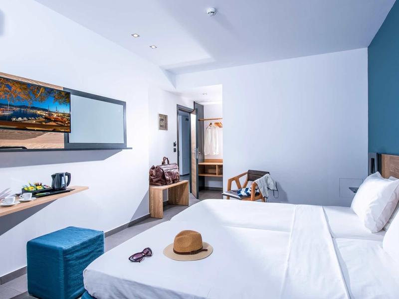 Modern hotel room with double bed, blue stool, desk, and open wardrobe.