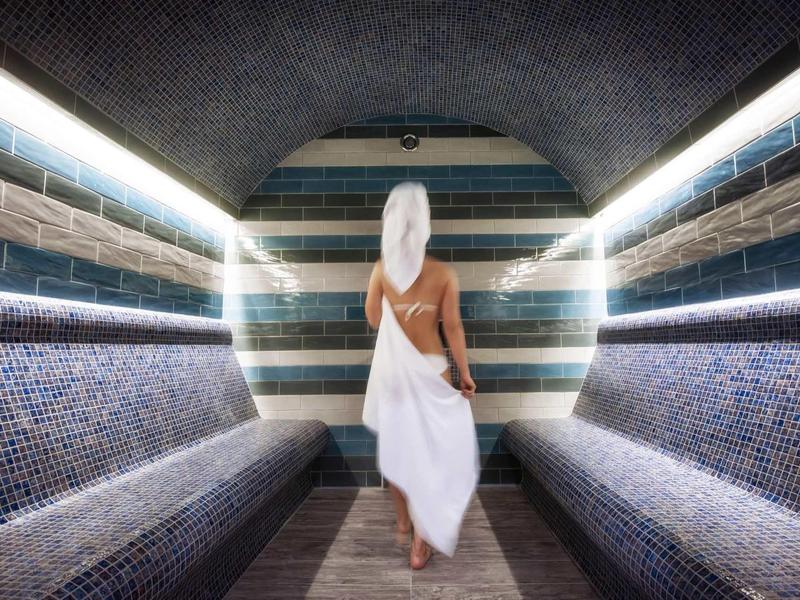 Woman in towel walking in steam room with mosaic walls and benches.