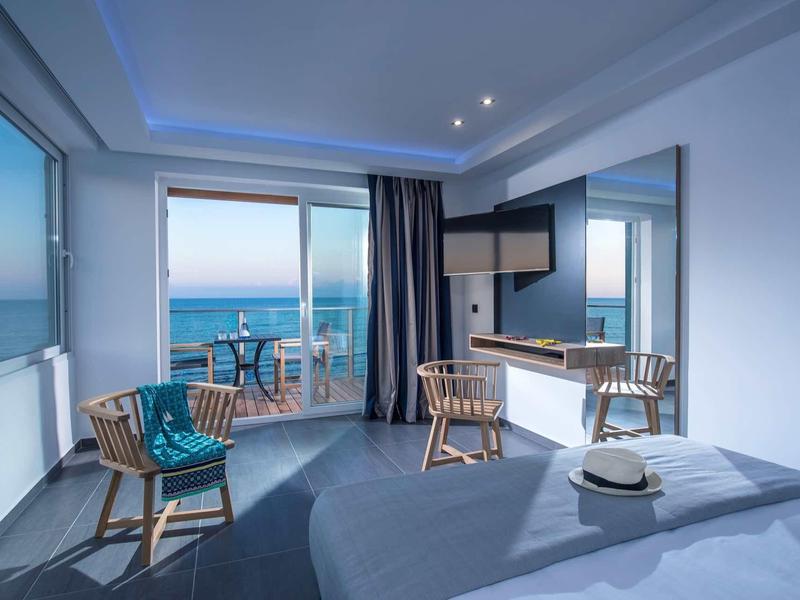 Bright hotel room with sea view, two chairs, and terrace at sunset.