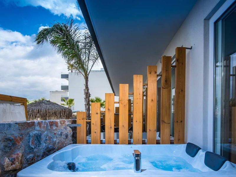 Modern hot tub on a terrace beside a wooden privacy screen and palm trees under a blue sky.