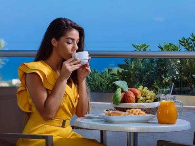 Frau in gelbem Kleid trinkt Kaffee auf Balkon mit Meerblick, Tisch mit Frühstück und Orangensaft.