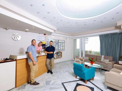 Drei Männer stehen in modernem Wohnzimmer mit hellen Wänden, blauen Sesseln und großem Deckenlicht.