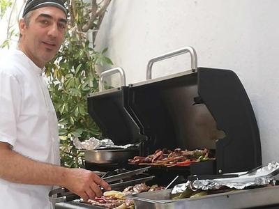 Mann grillt Fleisch auf einem Grill vor einer weißen Hauswand im Freien.