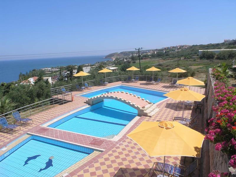 Schwimmbad mit Brücke, gelben Sonnenschirmen und Blick aufs blaue Meer im Hintergrund.
