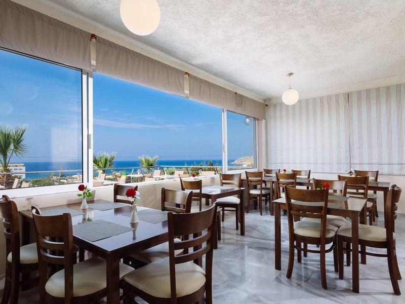 Restaurant mit Holztischen und -stühlen, großen Fenstern und Blick auf das blaue Meer.