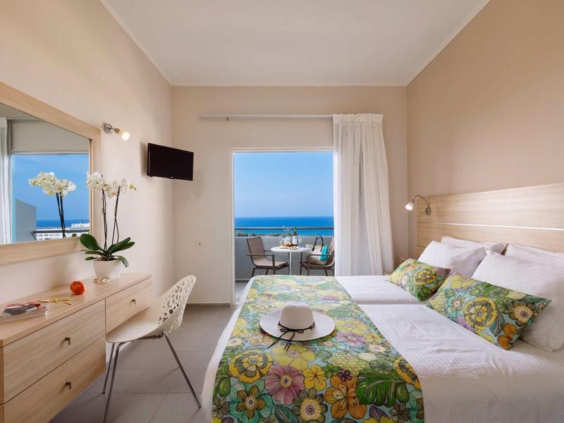 Helles Schlafzimmer mit Bett, buntem Bettläufer, Schreibtisch, Orchidee und Meerblick durch große Tür.