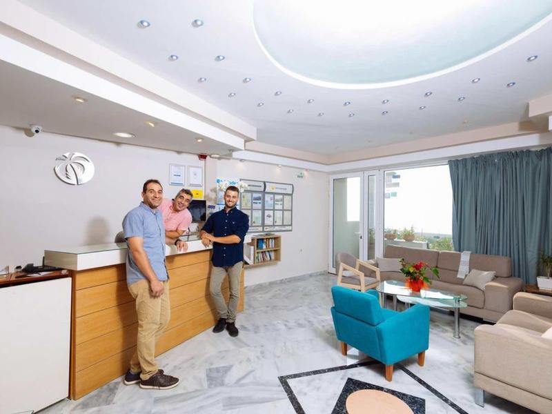 Drei Männer stehen in modernem Wohnzimmer mit hellen Wänden, blauen Sesseln und großem Deckenlicht.