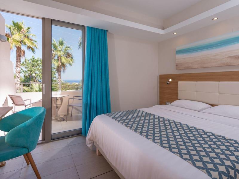 Jasny pokój hotelowy z podwójnym łóżkiem, niebieskim krzesłem i balkonem z palmami.