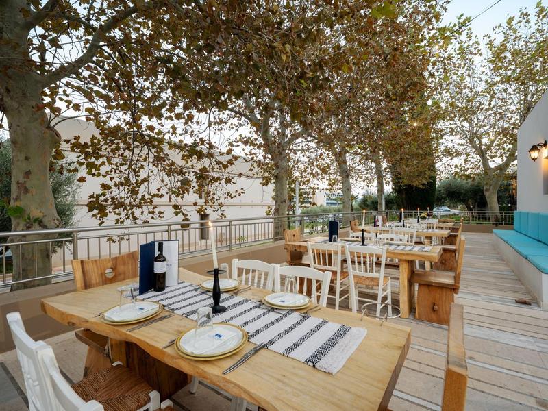 Przytulna zewnętrzna część restauracji z drewnianymi stołami i białymi krzesłami pod drzewami.